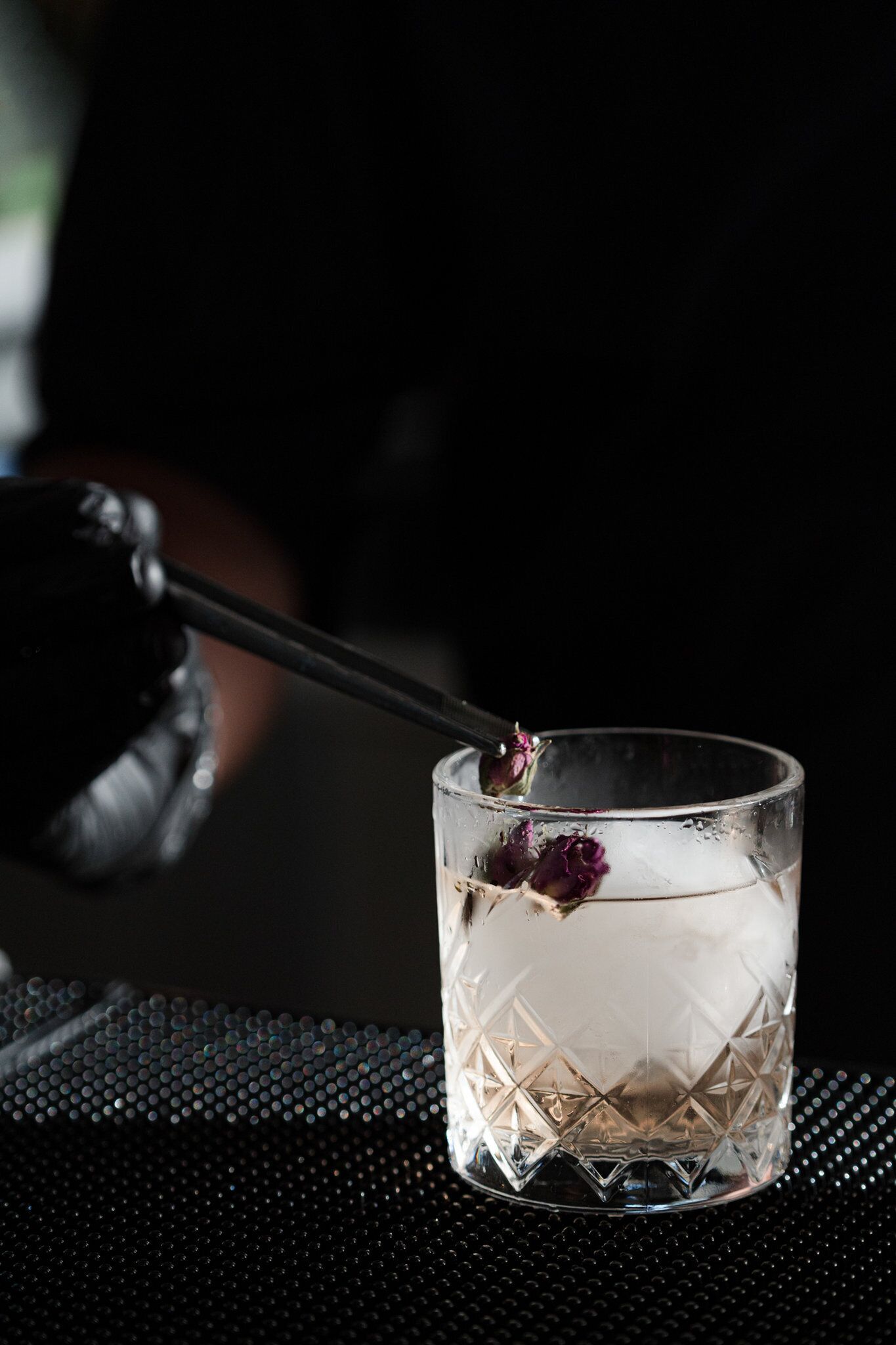 Un verre transparent orné d’un motif géométrique, contenant un liquide foncé et une fleur violette.