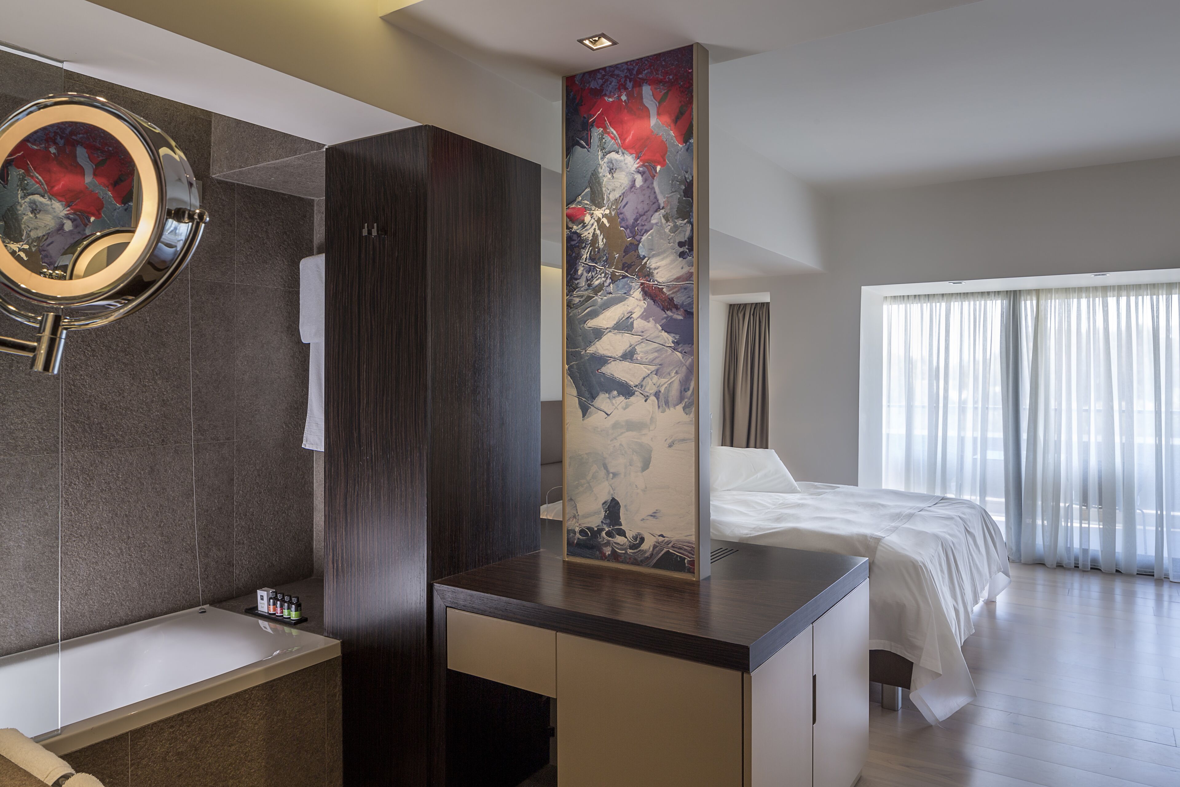 A modern hotel room with a large bed, a built-in desk, a bathroom with a bathtub, and a large abstract painting on the wall.