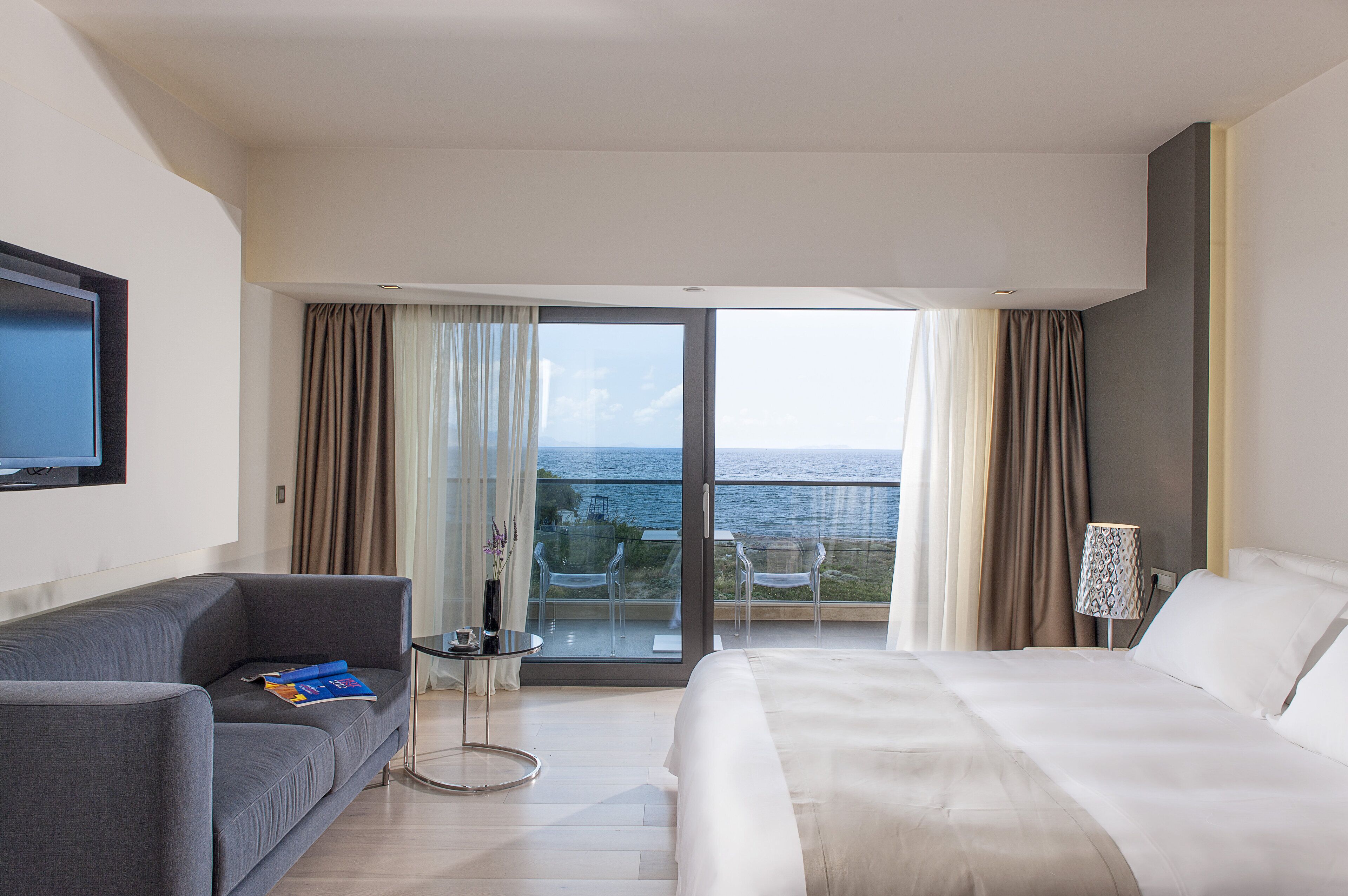 A hotel room with a bed, a sofa, a TV, a balcony with a view of the sea, and a table with a lamp.
