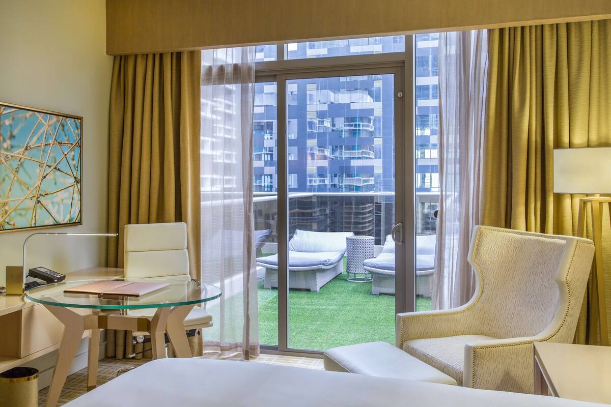 Habitación de hotel con una mesa de vidrio, una silla, una cama, un escritorio con lámpara, un cuadro y vistas a un balcón con mobiliario exterior.