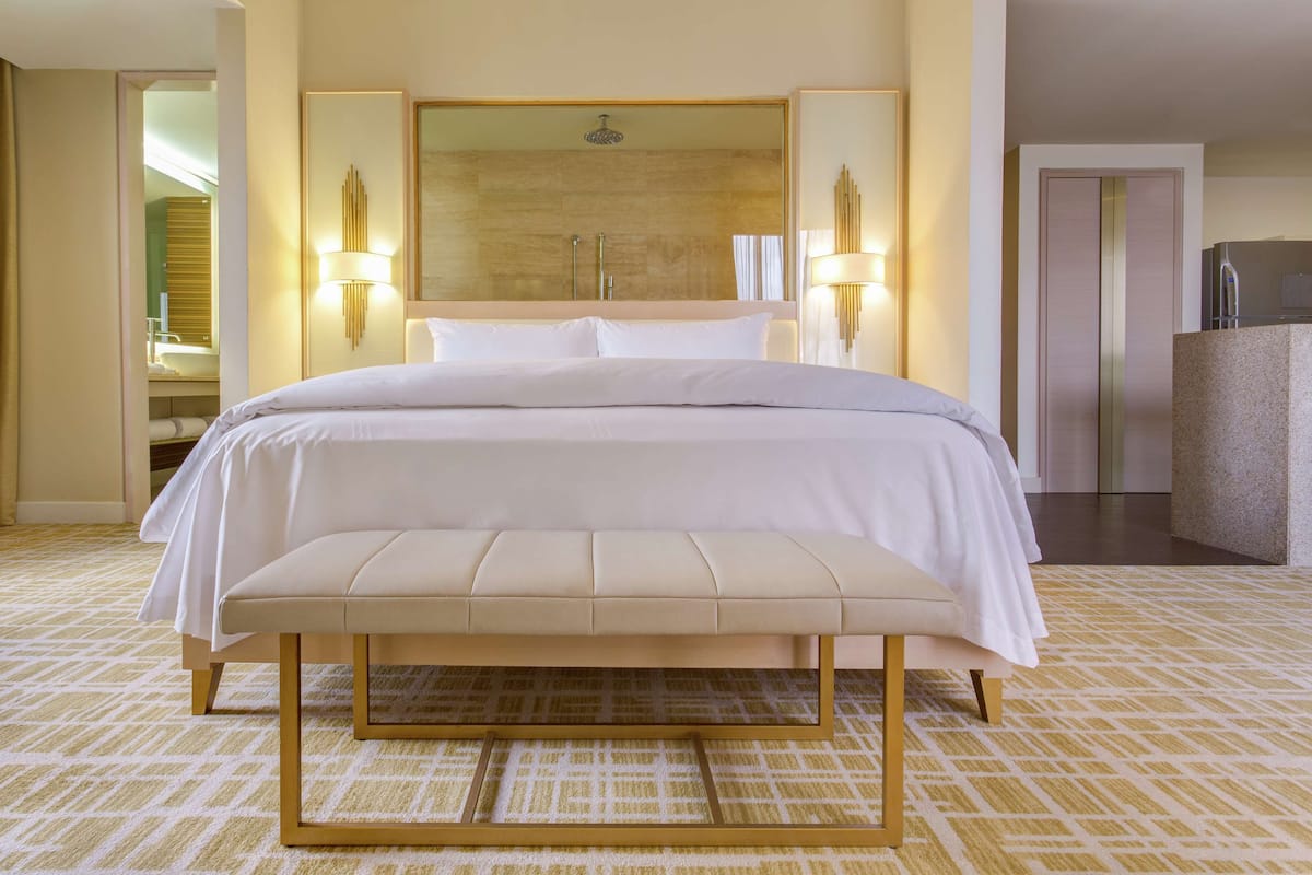 Habitación de hotel con una cama grande, un banco, un espejo y un baño visible a través de una puerta abierta.