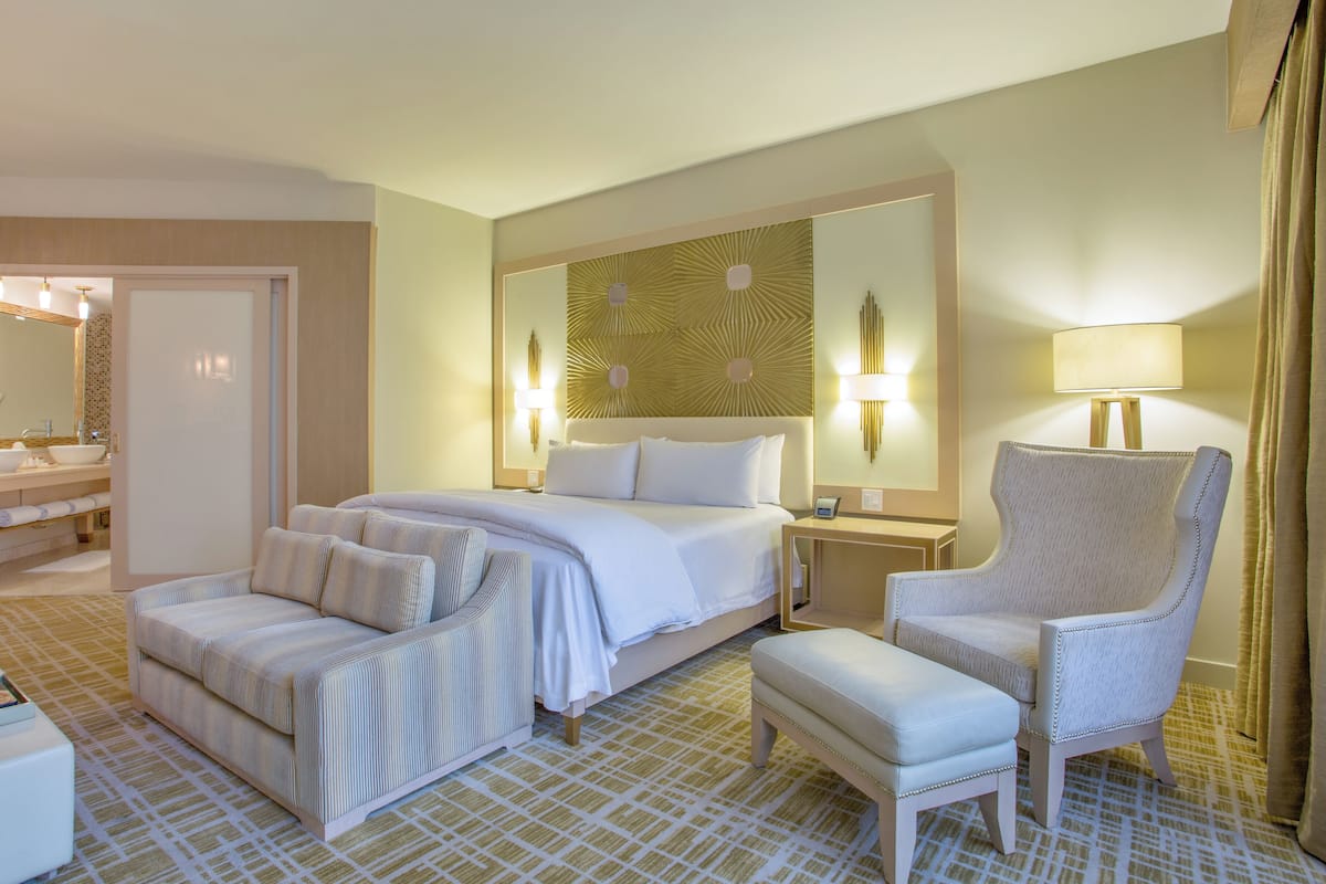 Habitación de hotel con cama, una silla, un banco, una mesita de noche y un baño visible a través de una puerta abierta.