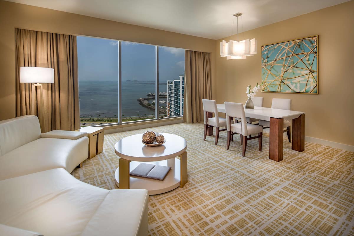 Habitación de hotel con un ventanal que ofrece vistas al mar, un comedor con mesa y sillas, y un sofá.