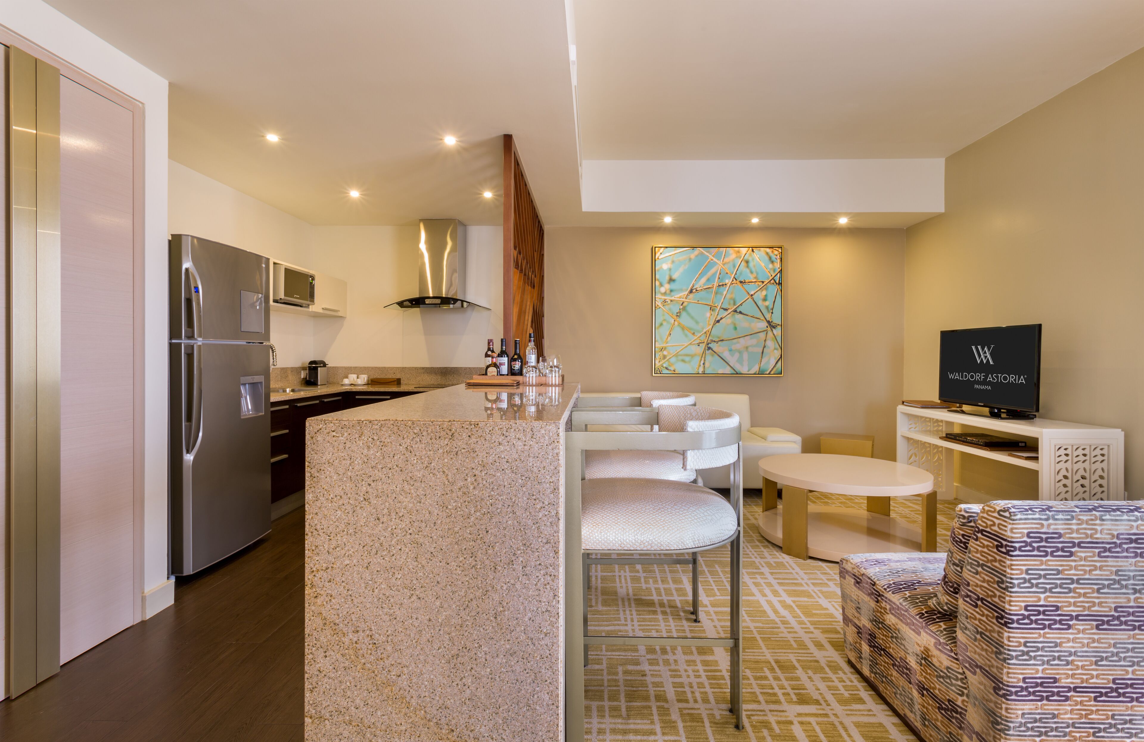 A modern kitchen with a built-in refrigerator, microwave, and stove. There is a dining area with a white table and chairs, and a patterned sofa. A television is mounted on the wall.