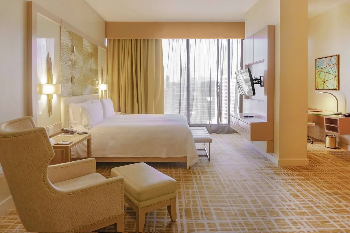 Una habitación de hotel con una cama grande, una zona de estar con una butaca y reposapiés, un escritorio con computadora y televisión.