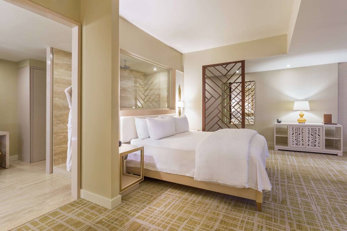 Habitación de hotel con cama, mesita de noche con lámpara, espejo, baño con ducha y una banca.