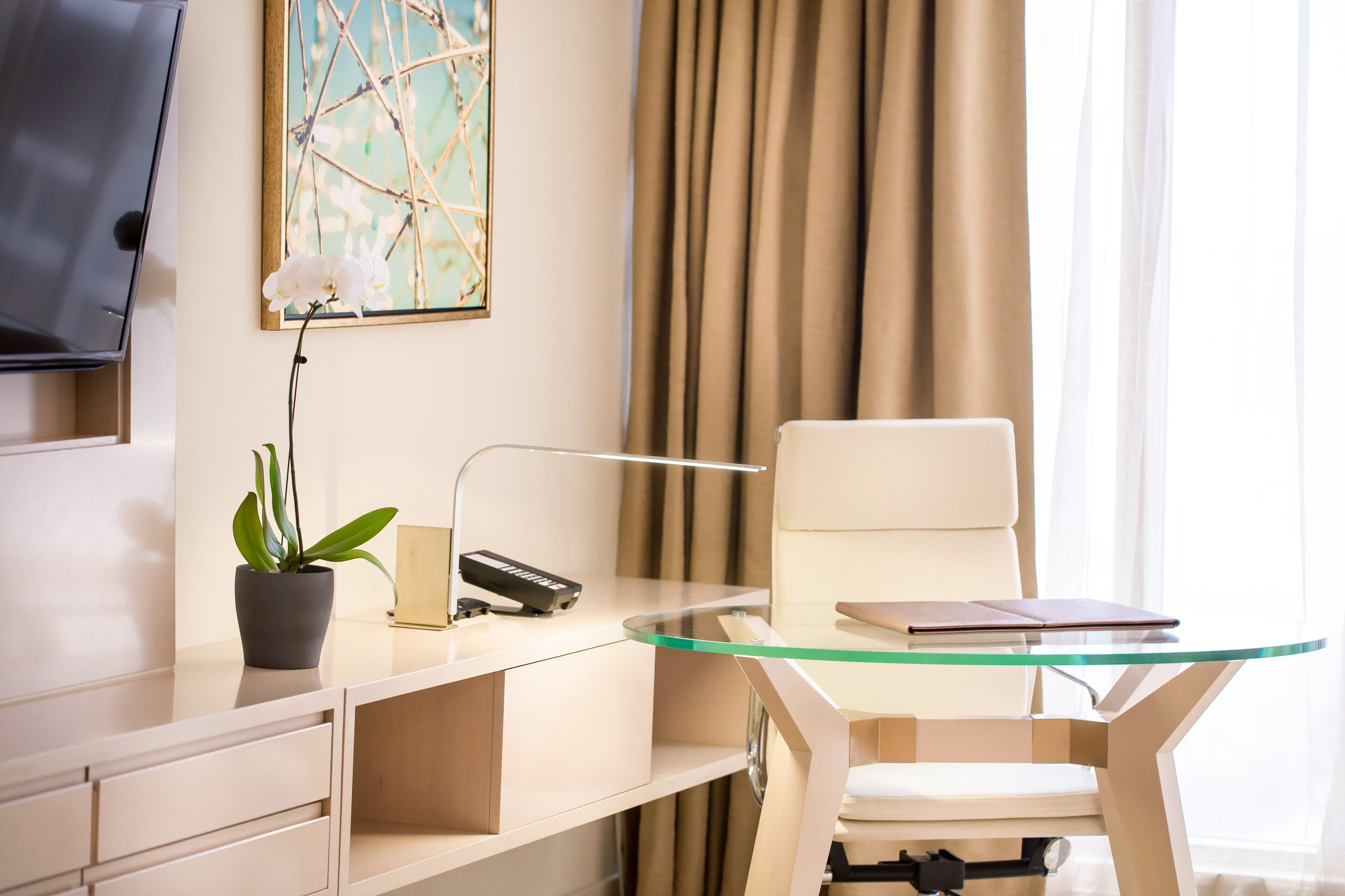 Un hotel moderno con un escritorio de vidrio, una silla blanca, una planta en maceta, un televisor de pantalla plana y un cuadro en la pared.