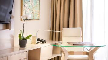 Un hotel moderno con un escritorio de vidrio, una silla blanca, una planta en maceta, un televisor de pantalla plana y un cuadro en la pared.