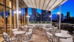 Un espacio al aire libre con sillas y mesas de mimbre, amplias ventanas que ofrecen vistas de la ciudad al atardecer.