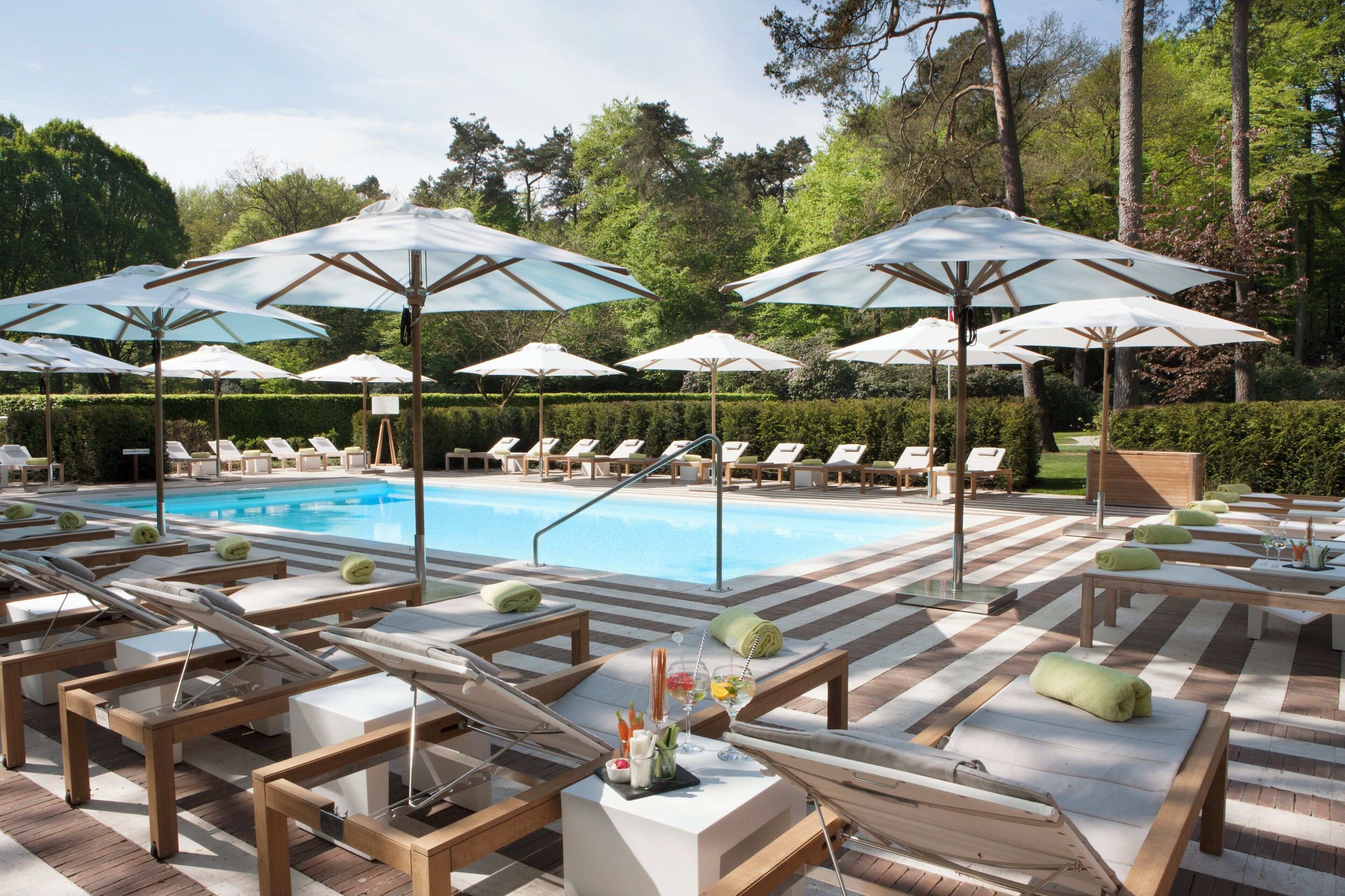 A poolside area with lounge chairs and umbrellas, surrounded by trees and a hedge.