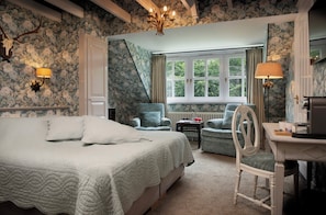 A bedroom with a large bed, two chairs, a desk, and a window with curtains.