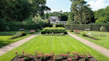 A well-manicured garden with a symmetrical layout, featuring a central hedge, flower beds, and a pathway leading to a large house.