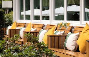 Outdoor seating with wooden benches and yellow cushions, arranged in front of a window with a view of umbrellas. - Landgoed Hotel Het Roode Koper (Ermelo)