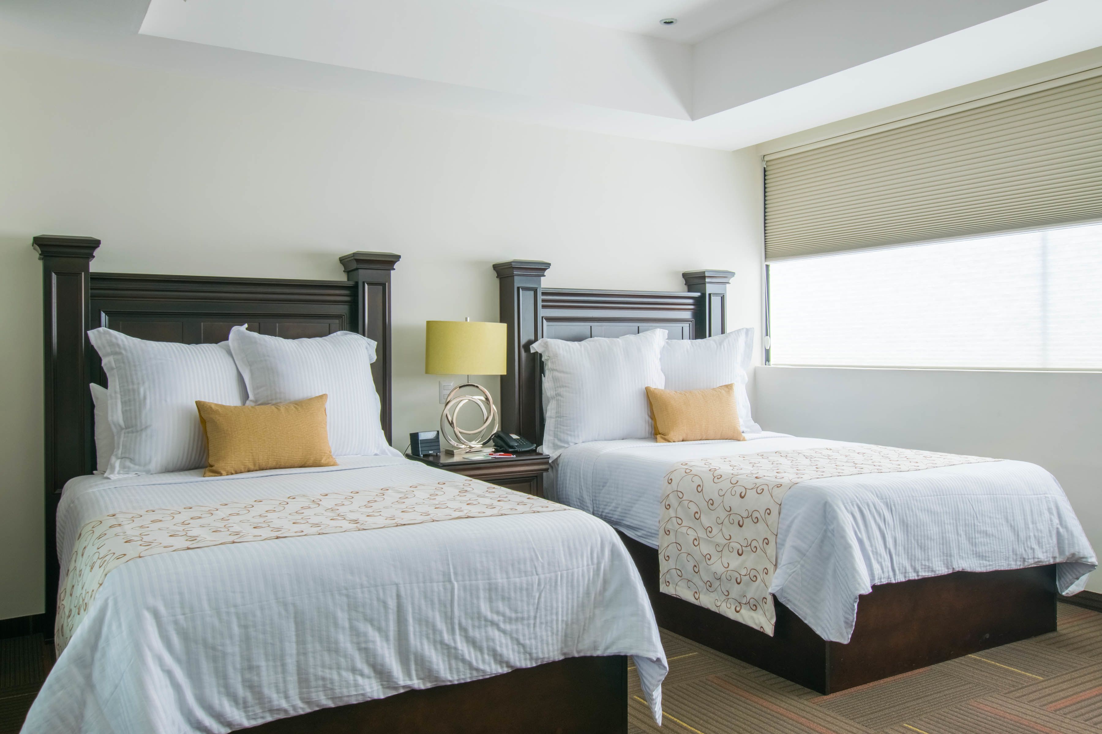 Habitación de hotel con dos camas, cada una con cabecera de madera, ropa de cama blanca y una sola almohada amarilla. Hay una mesita de noche con una lámpara y un teléfono.