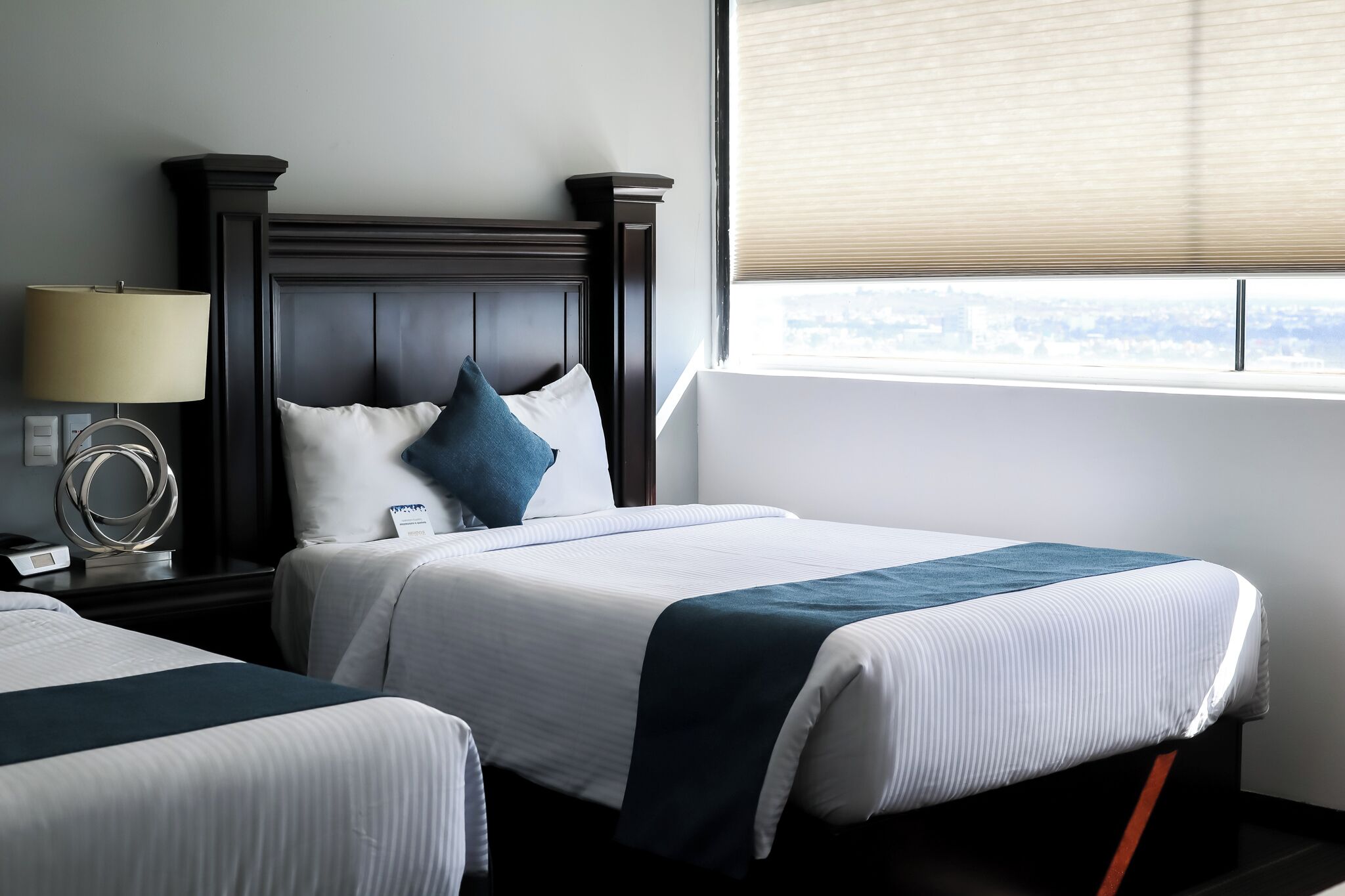 Une chambre d’hôtel avec deux lits, une table de chevet avec une lampe et une fenêtre avec des stores.