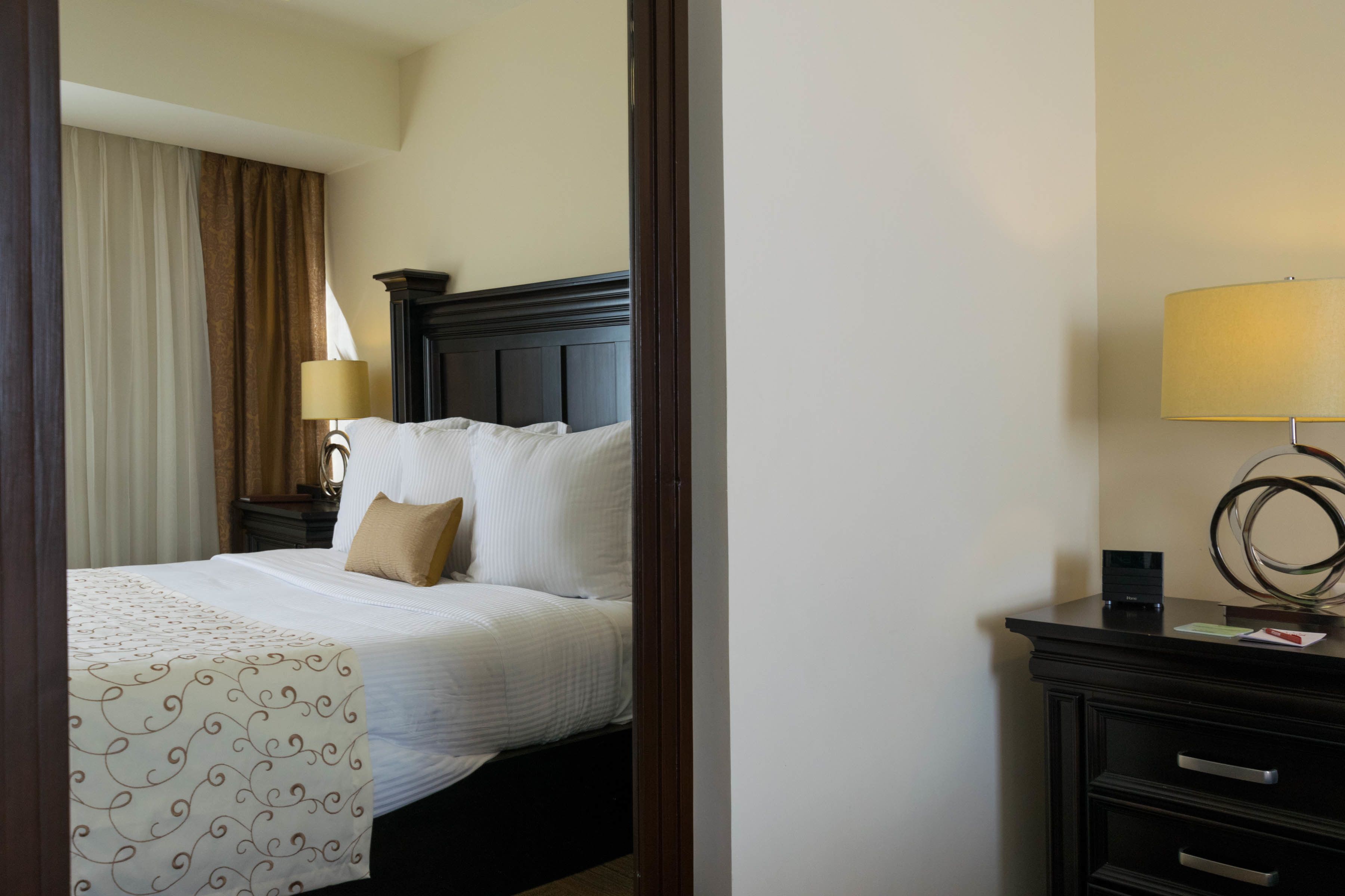 A hotel room with a neatly made bed, a bedside lamp, a dark wooden nightstand, and a wall-mounted lamp.