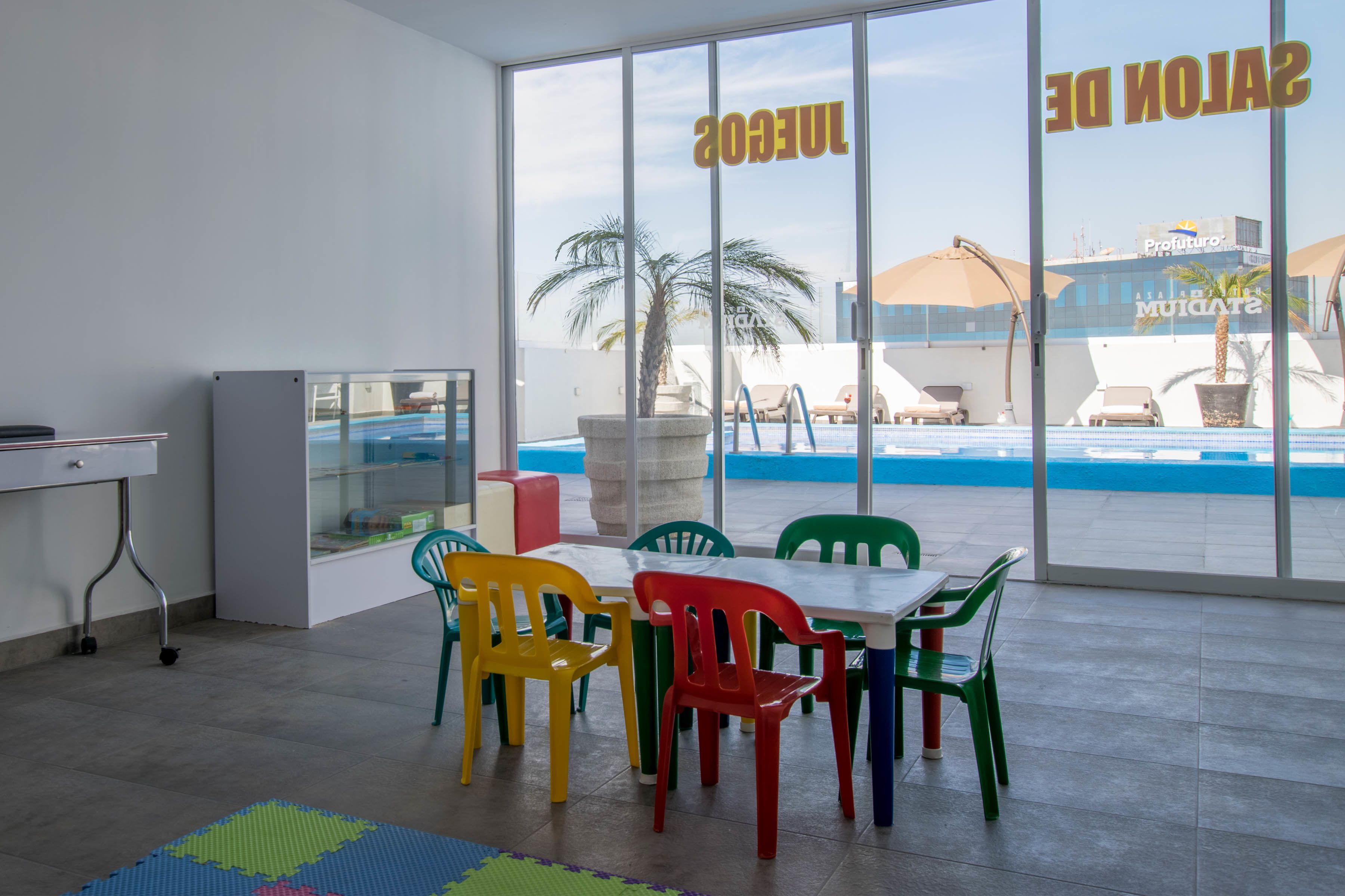 Una habitación con un ventanal grande que da a una piscina y vistas de un edificio con un letrero. Hay sillas coloridas y una mesa en la habitación.
