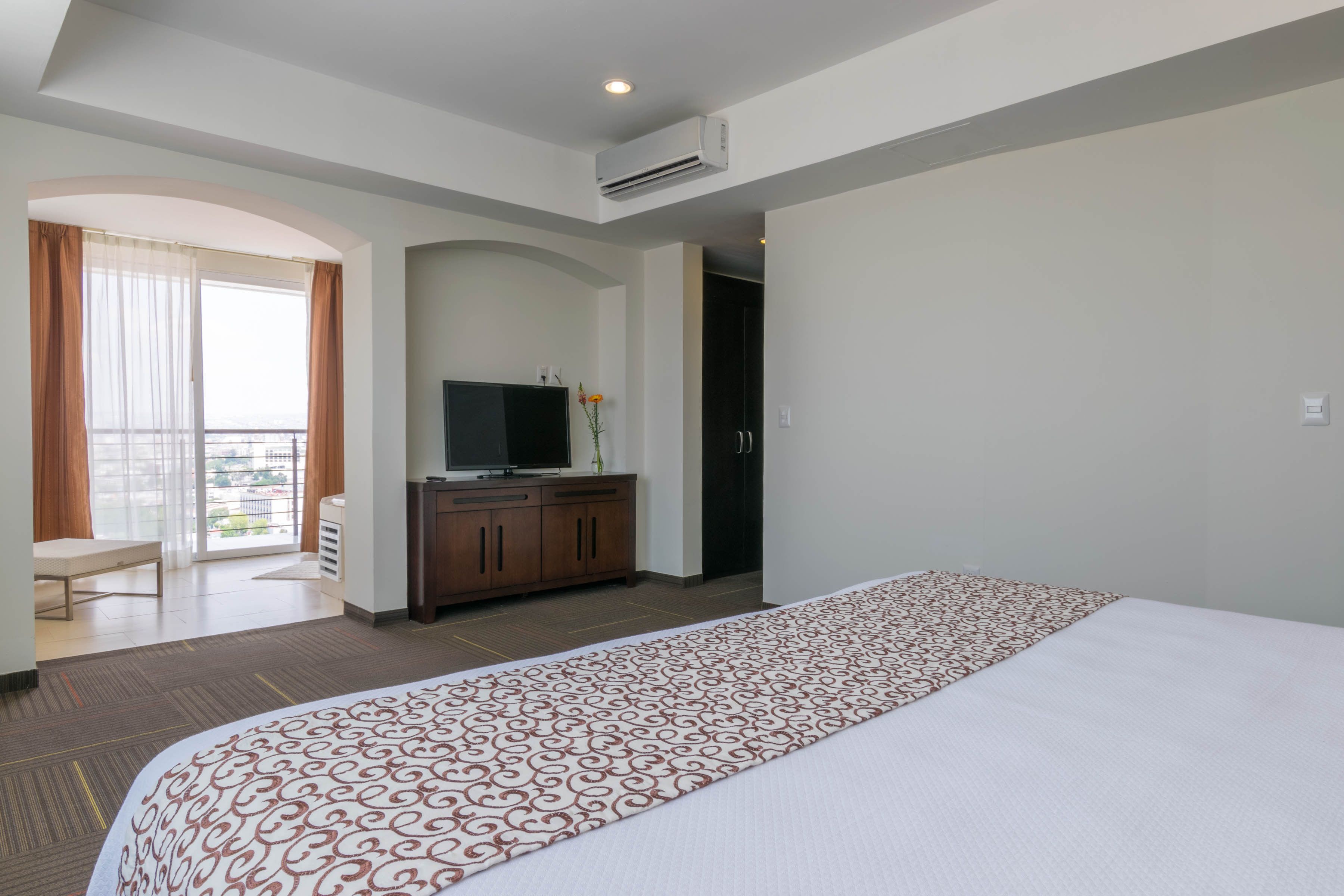 A hotel room with a large bed, a television on a wooden cabinet, a balcony with curtains, and a view of the city.
