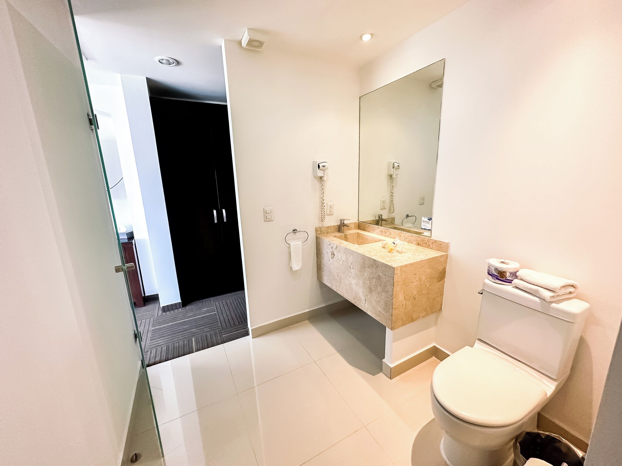 A modern bathroom with a glass door leading to a dark room, a marble sink, a large mirror, and a toilet.