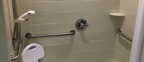 Une douche accessible aux personnes en fauteuil roulant, équipée d’une barre d’appui, de toilettes blanches et d’un siège de douche.