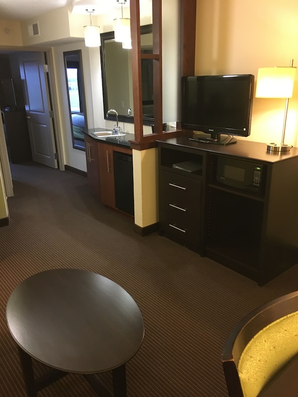 A hotel room with a kitchenette, a flat-screen TV, a microwave, a sink, a mirror, a dining table, and a chair. - AmericInn by Wyndham Fairfield (Fairfield)