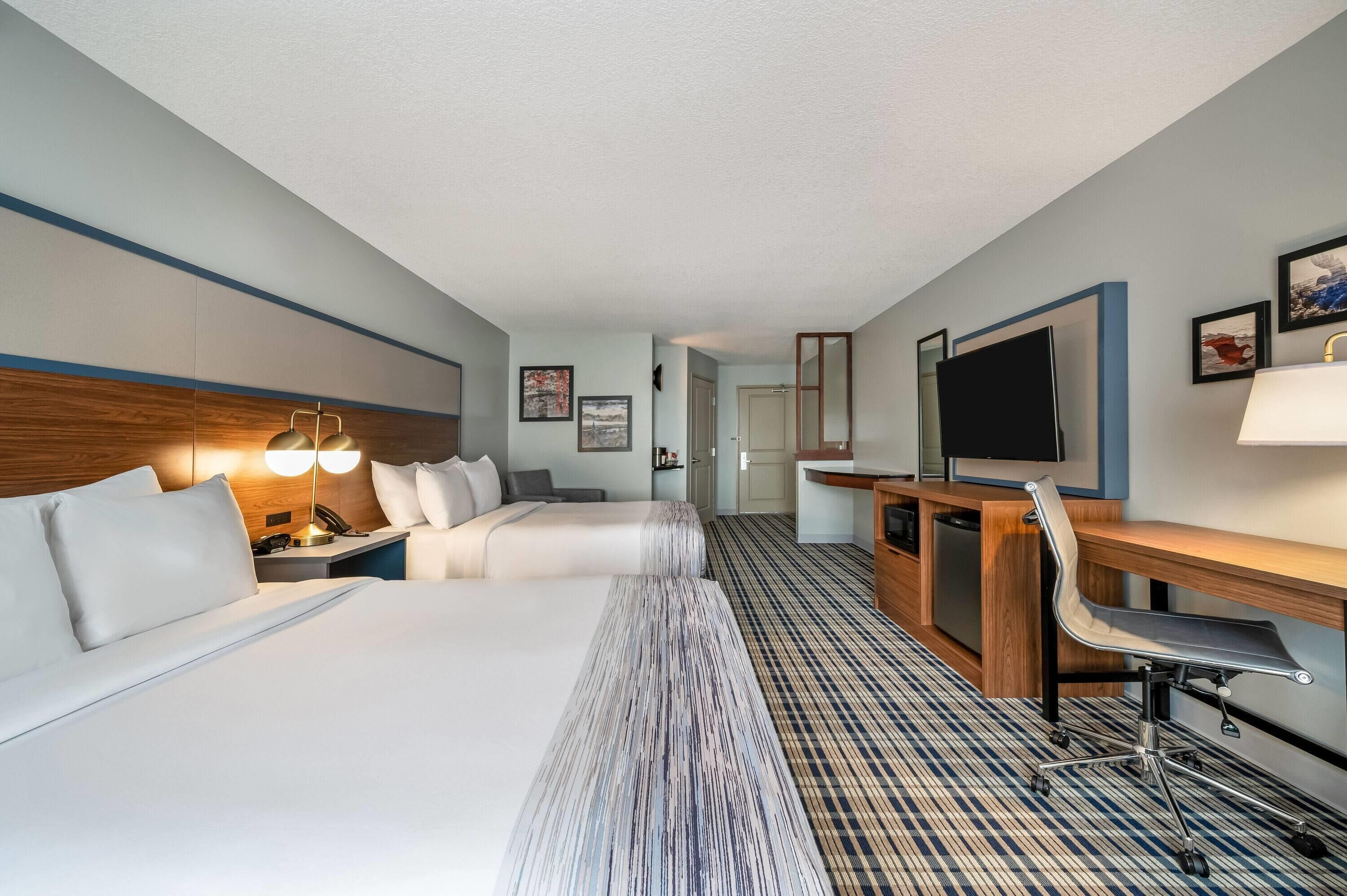 Quarto de hotel com duas camas, uma escrivaninha, uma cadeira, uma televisão e um abajur.