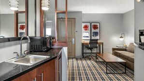 A compact hotel room with a kitchenette, a large mirror, a desk with a chair, a sofa, and a patterned rug. - AmericInn by Wyndham Fairfield (Fairfield)