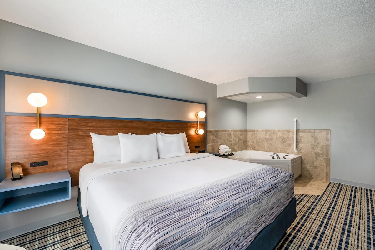 A hotel room with a large bed, a bedside table, a wall-mounted light, a built-in shelf, a bathroom with a bathtub, and a patterned rug.