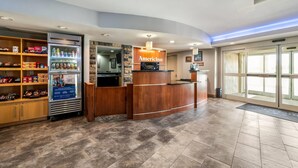 A reception area with a wooden counter, a refrigerator stocked with beverages, snack shelves, and a view of the outside through glass doors. - AmericInn by Wyndham Fairfield (Fairfield)