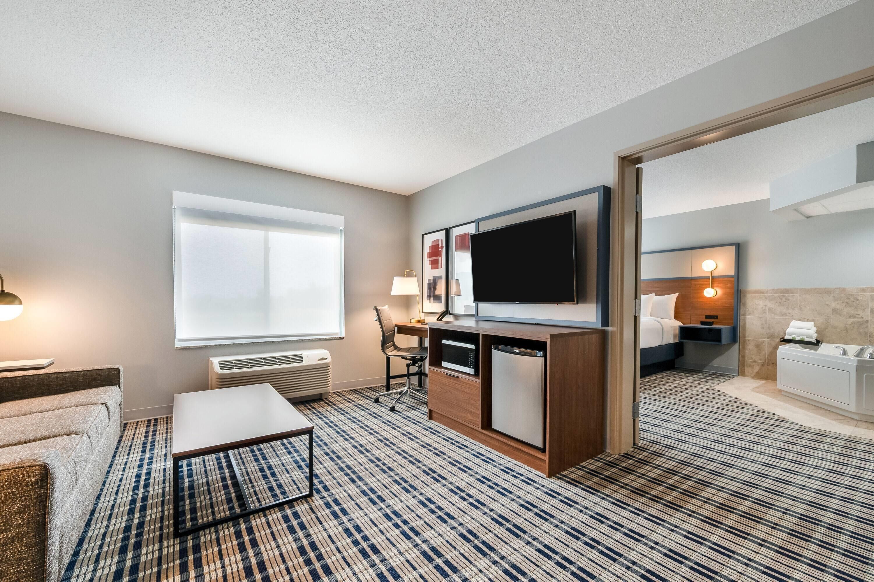 Ein Hotelzimmer mit Flachbildfernseher, Schreibtisch, Stuhl, Sofa, Couchtisch, großem Fenster, Spiegel und einem Badezimmer mit Waschbecken und Badewanne, sowie einem gemusterten Teppich.