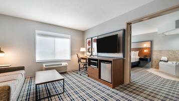 Ein Hotelzimmer mit Flachbildfernseher, Schreibtisch, Stuhl, Sofa, Couchtisch, großem Fenster, Spiegel und einem Badezimmer mit Waschbecken und Badewanne, sowie einem gemusterten Teppich.