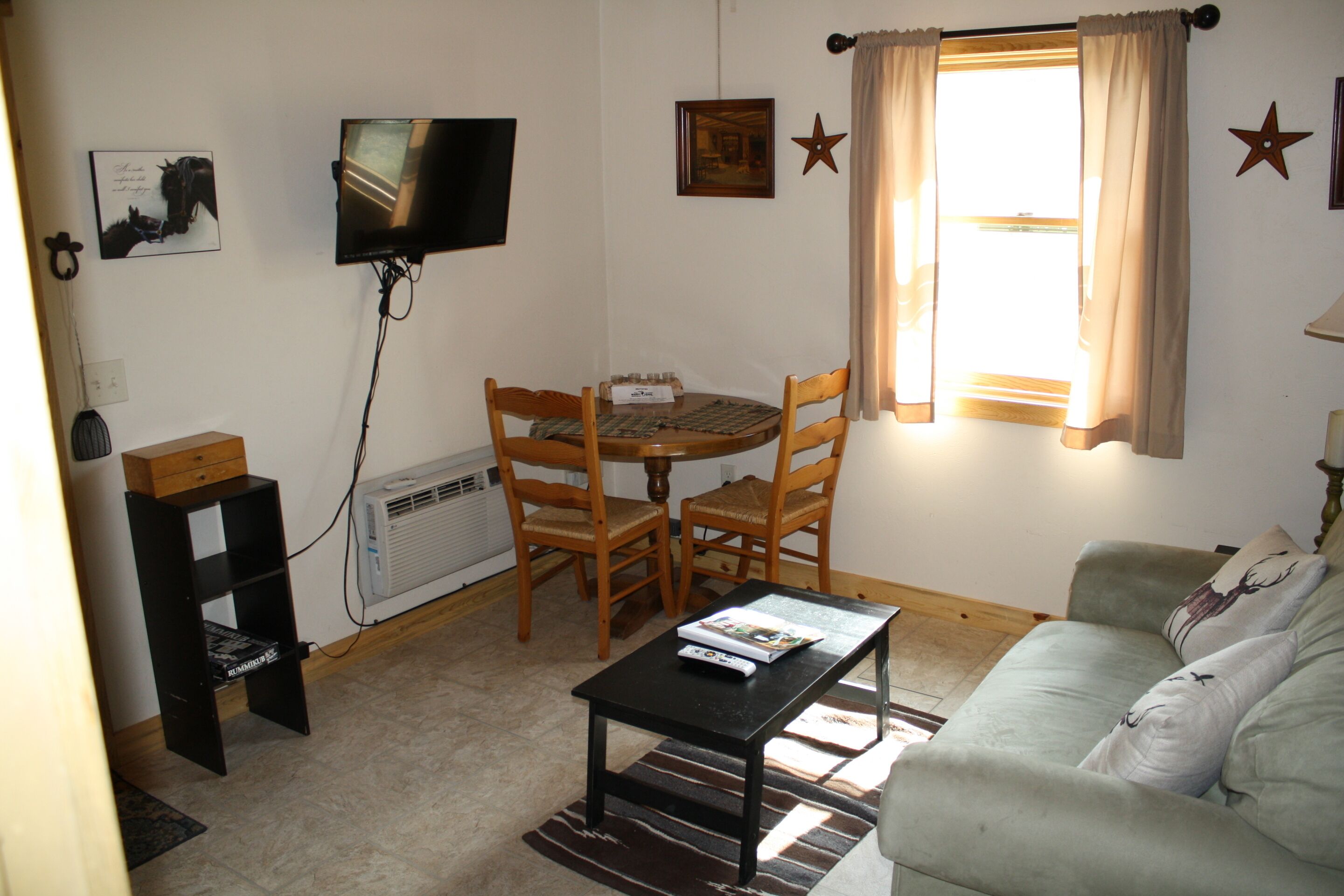 Uma sala de estar pequena com televisão, uma mesa, duas cadeiras, um sofá e uma janela com cortinas.