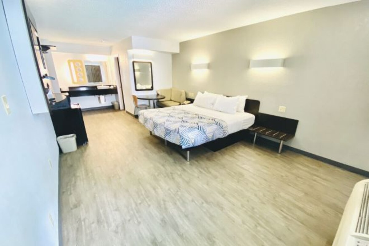 A hotel room with a bed, bedside table, and a small desk with a chair. There is a mirror on the wall and a light fixture above the bed.