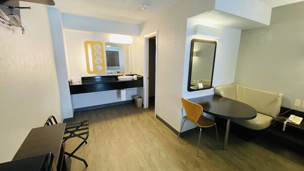 A modern hotel room with a wooden floor, a dining table, a sofa, a small desk, and a bathroom with a sink and mirror. - Motel 6 Boerne, TX (Boerne)