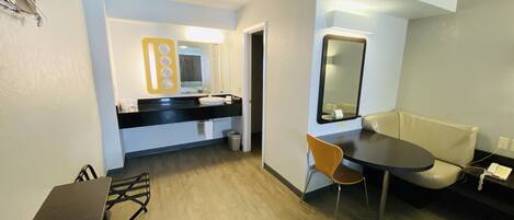 Une chambre d’hôtel moderne avec un plancher en bois, une table à manger, un canapé, un petit bureau et une salle de bain équipée d’un lavabo et d’un miroir.