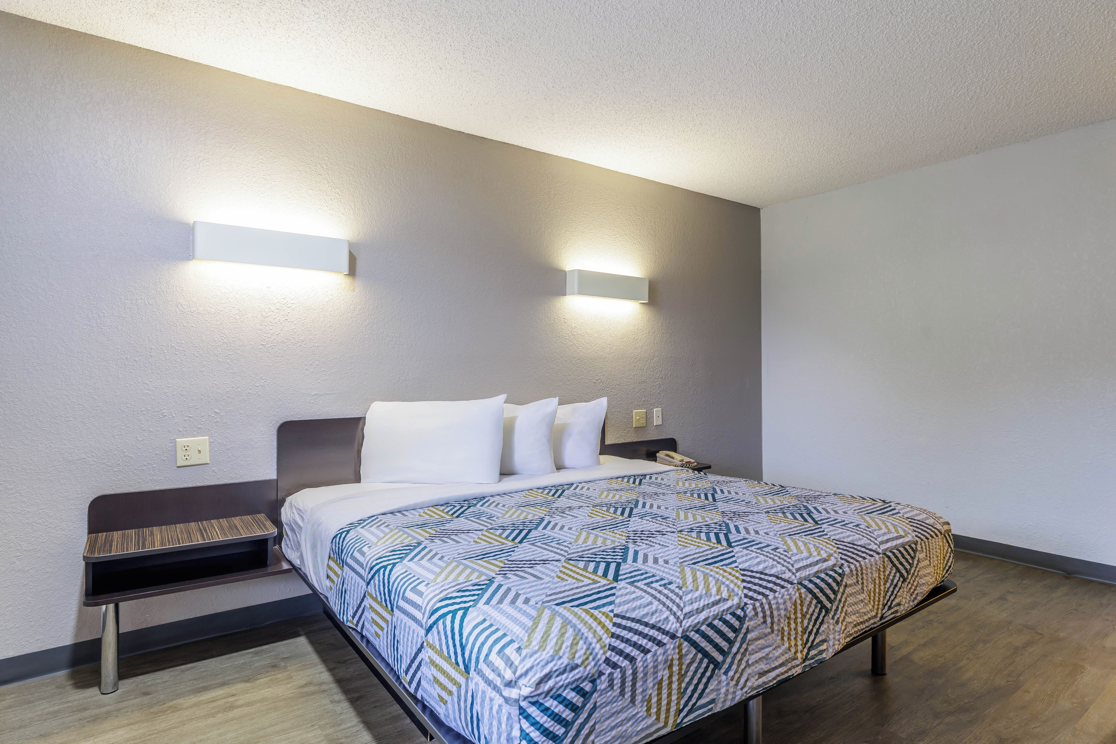 Una habitación de hotel con cama, dos mesitas de noche, colcha estampada e iluminación en la pared.