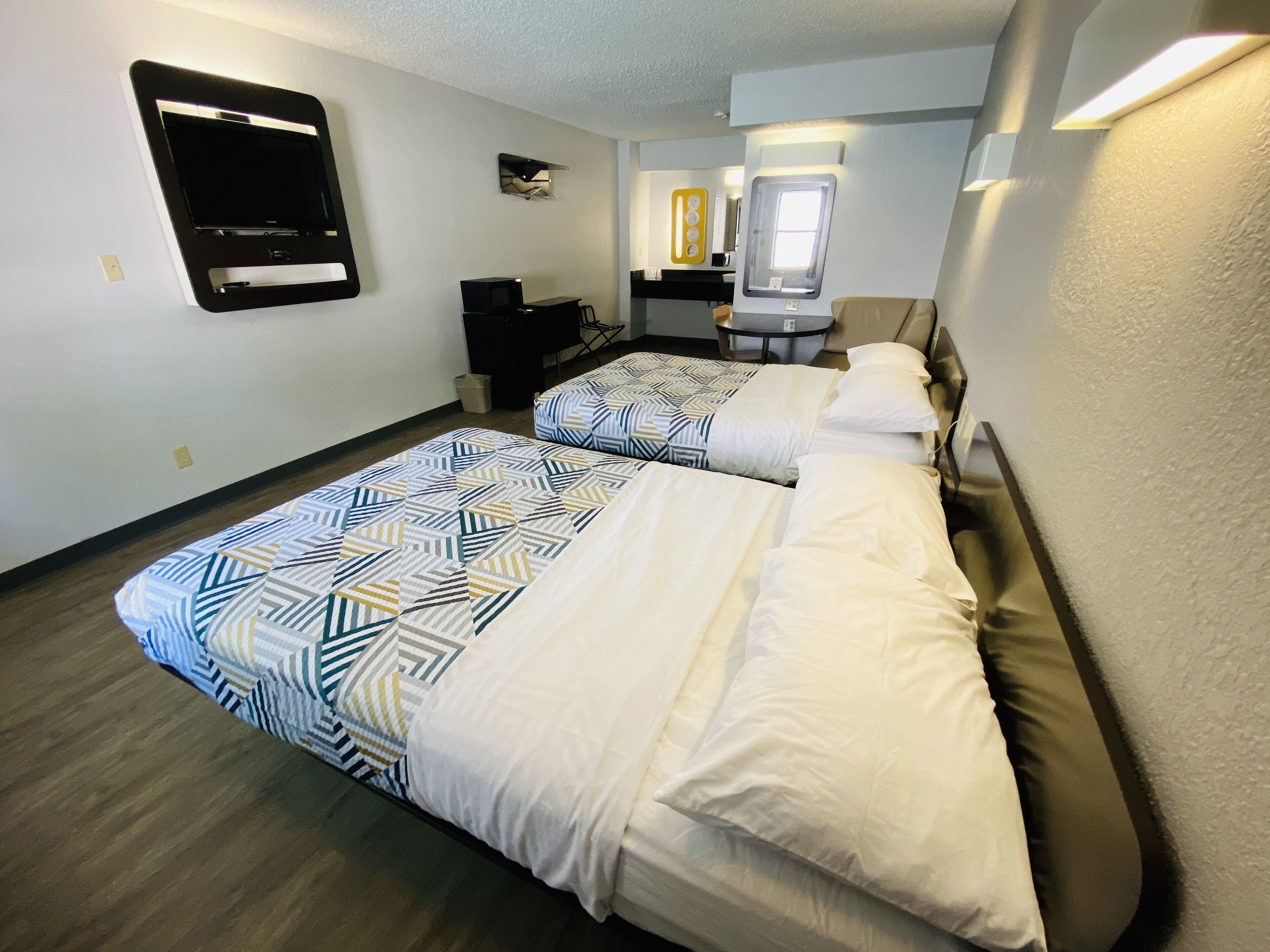 A hotel room with two beds, a small desk, a chair, and a window.