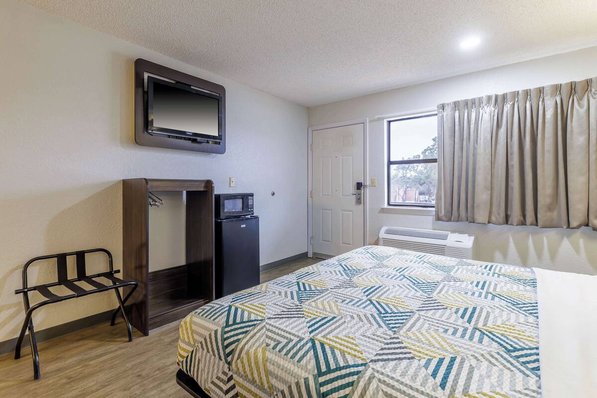 A hotel room with a bed, a TV mounted on the wall, a desk with a microwave, and a window with curtains.