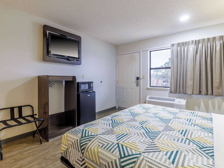A hotel room with a bed, a TV mounted on the wall, a desk with a microwave, and a window with curtains.