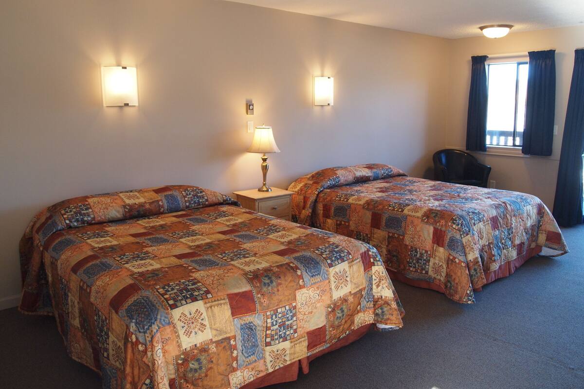 A hotel room with two beds, a nightstand with a lamp, a chair, and a window with curtains.