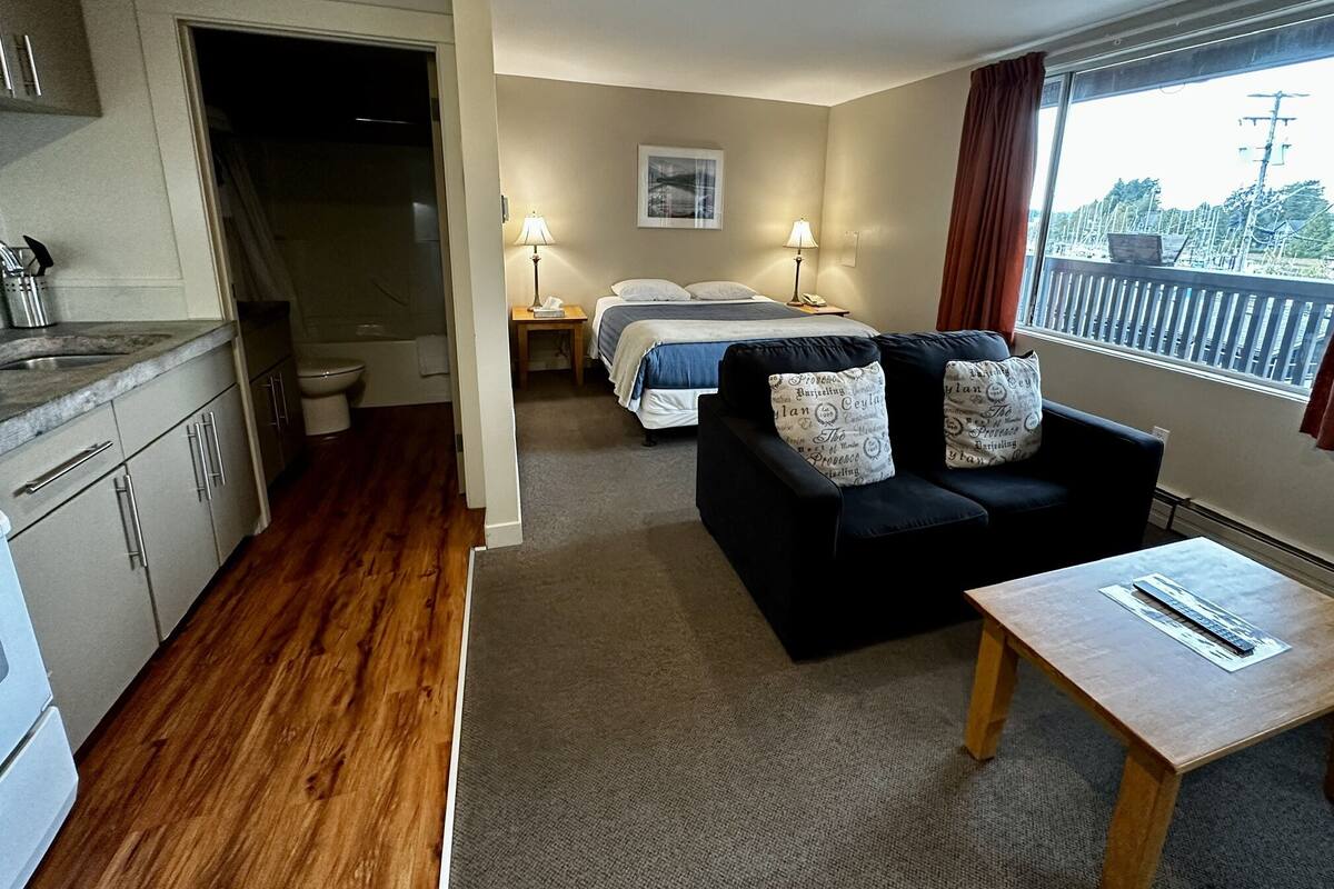A hotel room with a kitchenette, a bed with a blue blanket, a black sofa with patterned cushions, and a window with a view of trees and power lines.