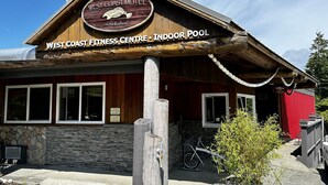 Exterior del West Coast Fitness Centre con piscina interior, destacando una fachada de madera, detalles de piedra y un letrero que indica que es un centro de acondicionamiento físico.