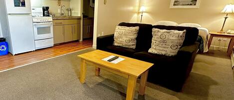 Una sala de estar con sofá, una mesa de centro, una zona de cocina con estufa y microondas, y un cuadro en la pared.