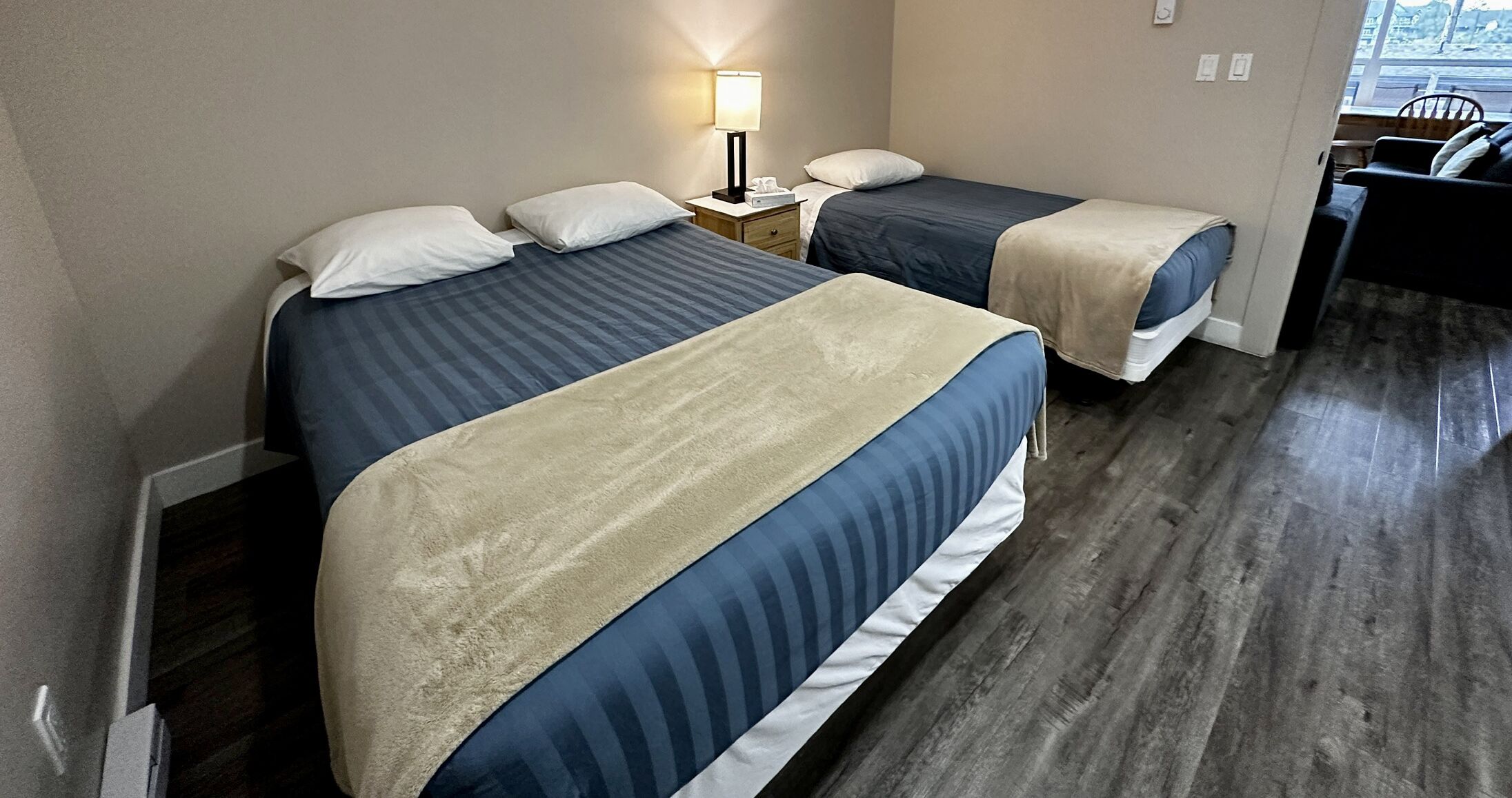 A hotel room with two beds, a nightstand with a lamp, and a view of the outside through a window.