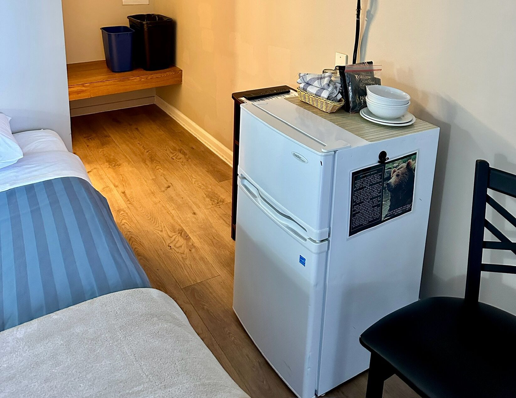 A compact room with a bed, a small refrigerator, a chair, and a shelf with items.