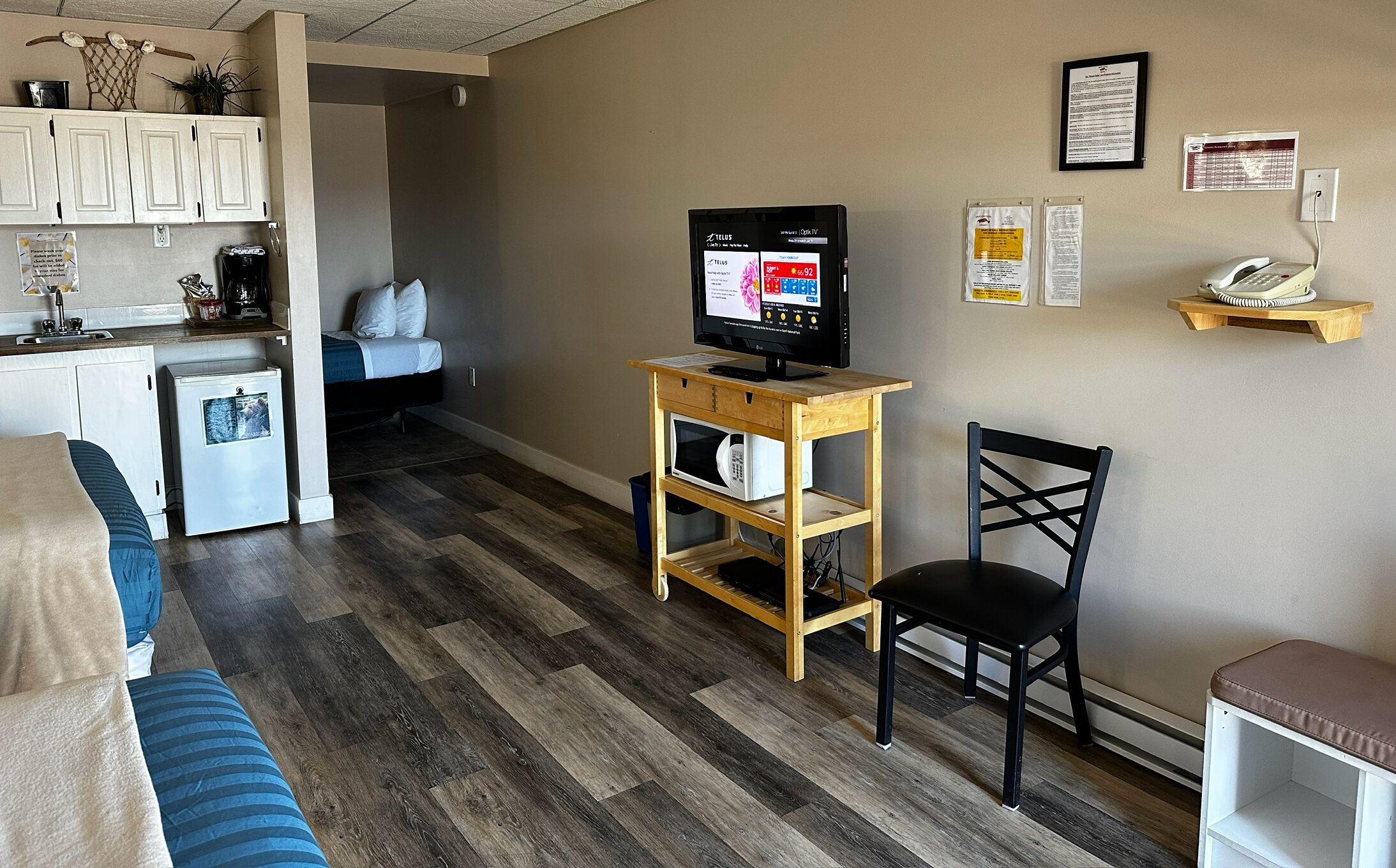 A compact living space with a kitchenette, a bed, a TV on a wooden stand, a chair, and a small desk.