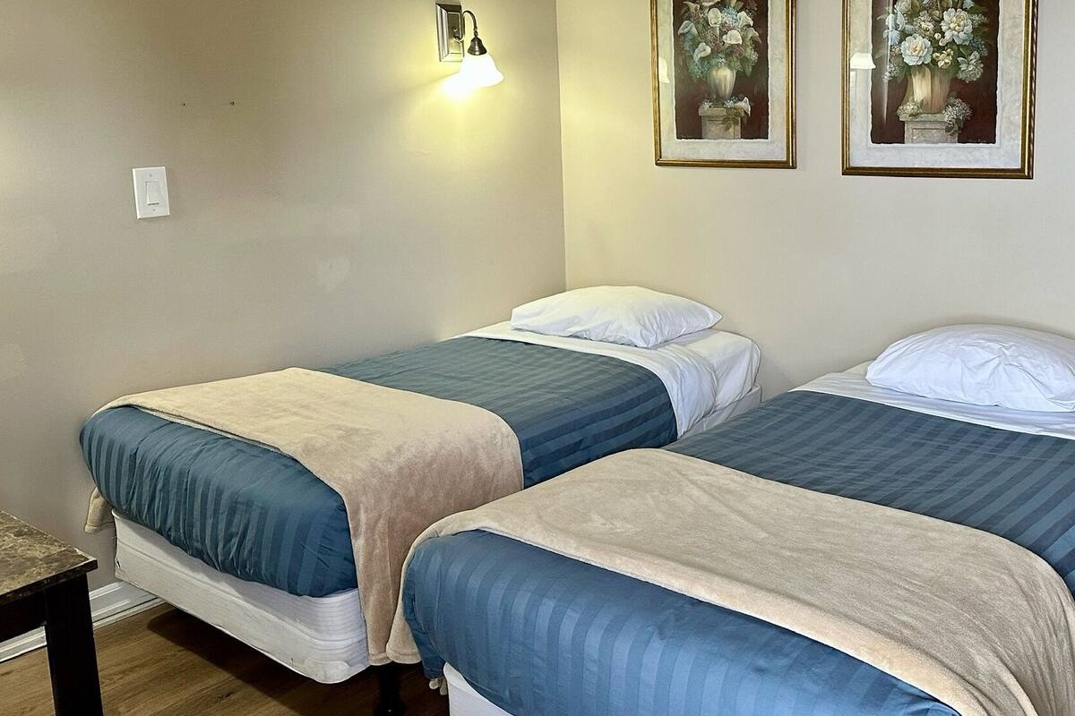 A room with two beds, each with a blue and beige bedspread, white pillows, and a wall-mounted light fixture. There are two framed floral paintings on the wall.