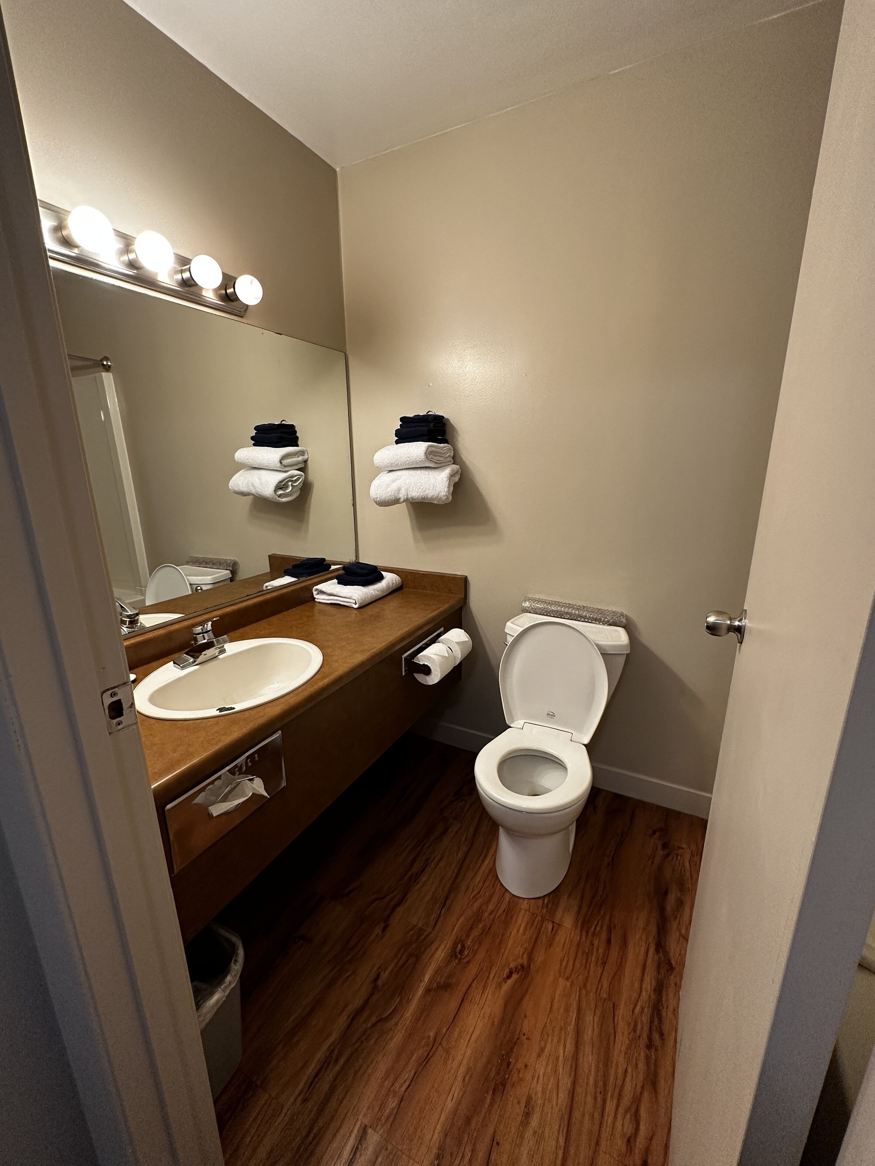 A bathroom with a wooden floor, a sink, a toilet, a mirror with lights, and towels on a rack.
