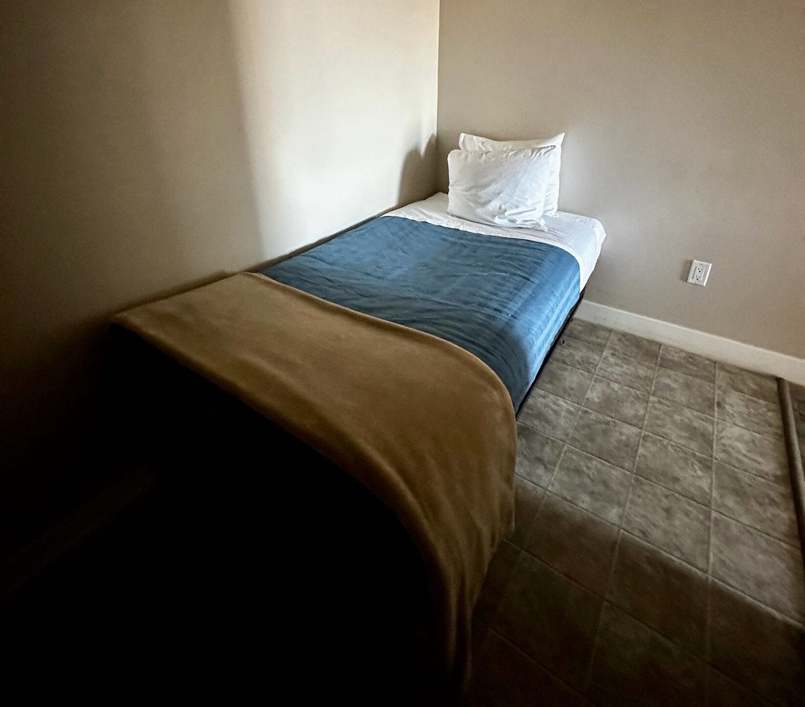 Quarto pequeno com cama de solteiro, edredom marrom e azul, travesseiros brancos e piso de azulejos.