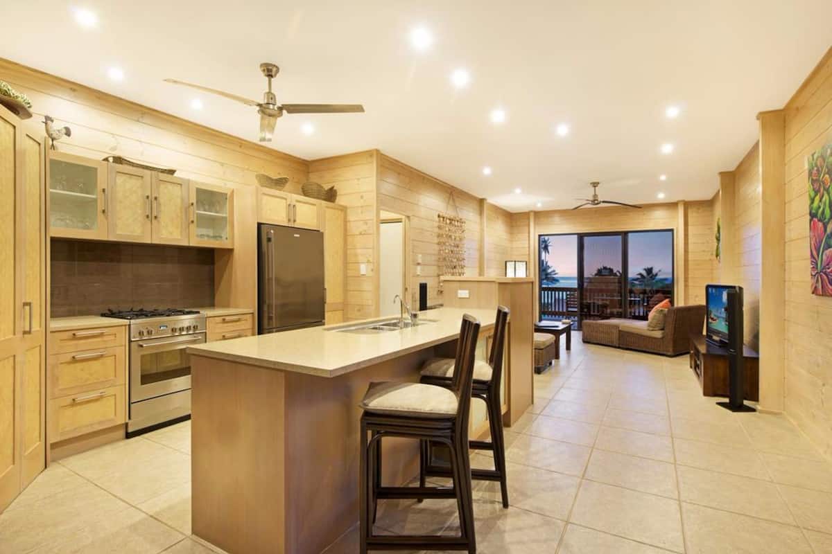 A modern kitchen with wooden cabinetry, a central island, and a dining area with a sofa and TV.